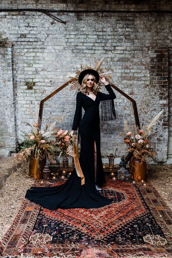a black A-line boho wedding dress with a depe neckline, fringe sleeves and a train plus a slit, silver boots and a black hat
