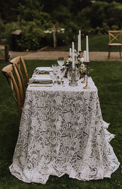 a boho lace wedding tablecloth looks incredibly chic and beautiful, tall and thin candles and greenery add chic to the table