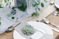 a chic wedding tablescape with a light grey table runner and napkin, greenery, succulents and elegant silver cutlery