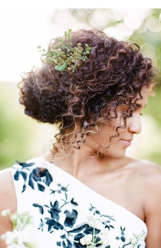 a curly low updo with some curls down and a fresh greenery branch tucked into the hairstyle for a fresh touch