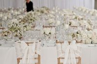 a refined white wedding reception space with lots of blooms and elegant bows wih lace on the couple’s chairs
