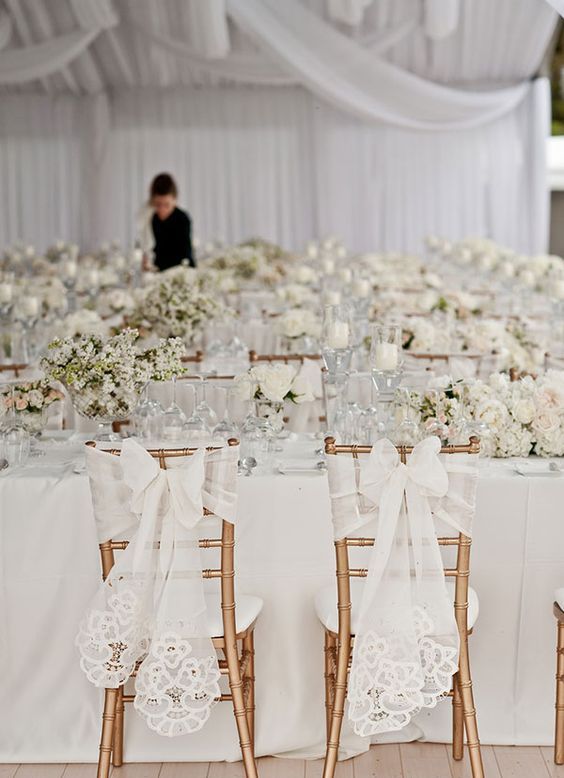 a refined white wedding reception space with lots of blooms and elegant bows wih lace on the couple's chairs