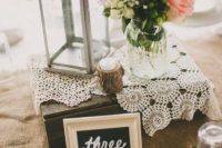 a rustic wedding centerpiece of a box, a couple of doilies, a branch candle holder, white and pink blooms and greenery and a candle lantern