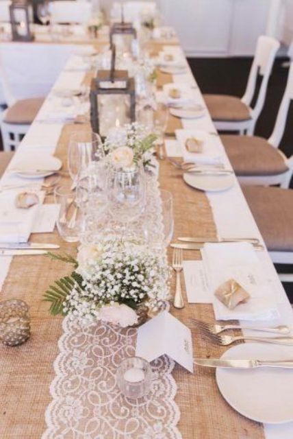 a rustic wedding tablescape with a burlap and lace table runner, white blooms and greenery and candle lanterns is fantastic