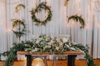 a sweetheart table with a lush greenery and pastel bloom table runner, lots of floating candles and wreaths with lights behind