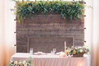 the sweetheart table accented with a wooden backdrop with greenery, candles, a crate with blooms and greenery