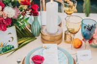 a bright and chic wedding tablescape with a blush tablecloth, pink napkins, yellow and blue glasses, red and pink blooms and greenery is a cool idea