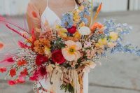 a cheerful wedding bouquet with orange,red, yellow and blue blooms, colorful feathers and tassels and fringe for a boho bride