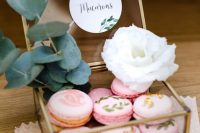 a clear glass and brass box with pink painted and gold leaf macarons, a white bloom and greenery is a beautiful idea to serve macarons at your wedding