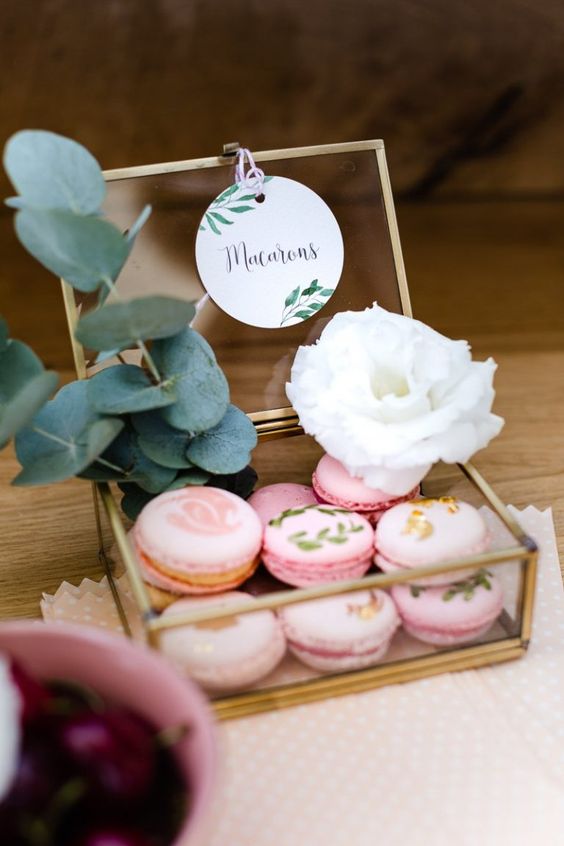 a clear glass and brass box with pink painted and gold leaf macarons, a white bloom and greenery is a beautiful idea to serve macarons at your wedding