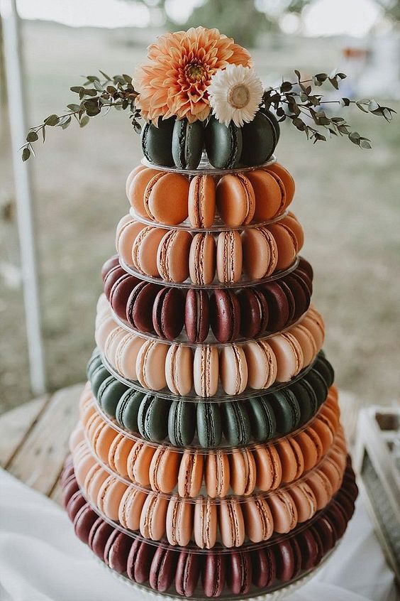 a clear multi-tier stand with lots of dark green, orange and burgundy macarons and some matching blooms and greenery on top