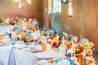 a colorful wedding table setting with blue, red, yellow and orange blooms, colorful candles, glasses and candleholders plus hanging bulbs