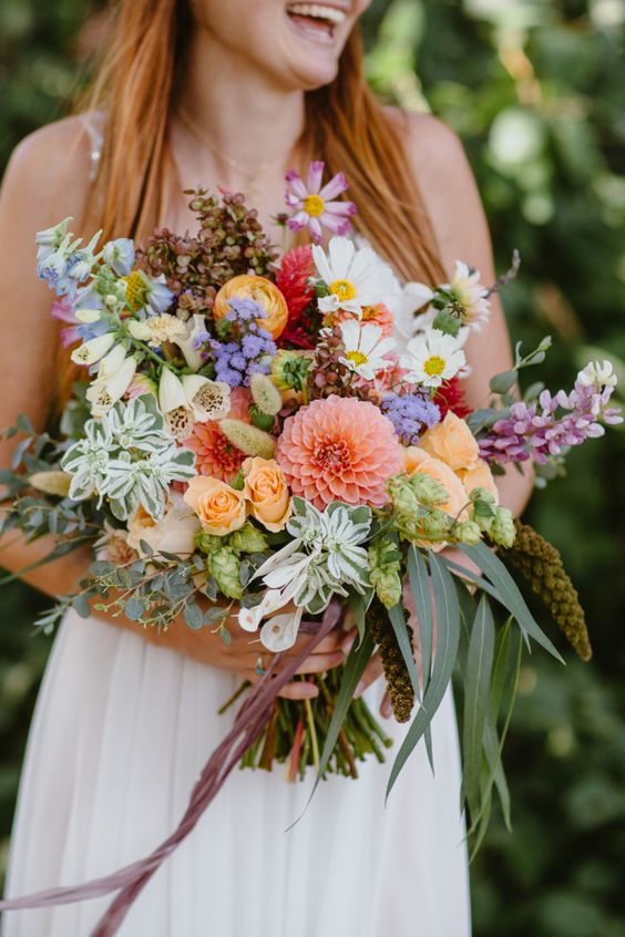 a hoppy summer wildflower wedding bouquet done in purple, white, yellow, orange and pink, with greenery and seed pods