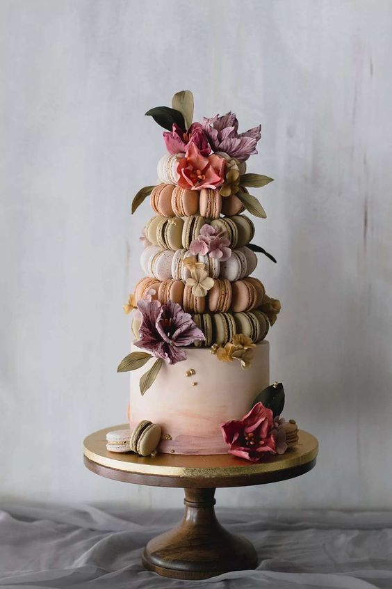 a watercolor wedding cake with a tower of macarons placed on top - orange, green and white ones, with greenery and pastel blooms