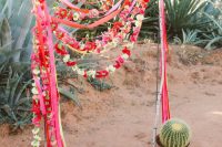 a Mexican fiesta wedding arch covered with bright blooms and ribbons and oversized potted cacti is bold and cool