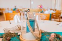 a beach wedding centerpiece with fishing net, seashells, a glass lantern with sand and a turquoise candle