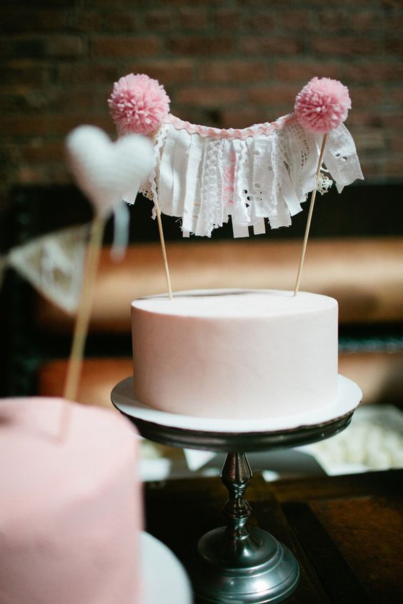 a blush buttercream wedding cake with a banner with lace pieces and pink pompoms is a gorgeous dessert for a wedding