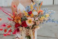 a cheerful wedding bouquet with orange,red, yellow and blue blooms, colorful feathers and tassels and fringe for a boho bride