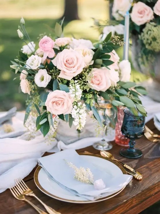 a chic pastel wedding tablescape with a pale blue table runner and napkins, blush roses and greenery, navy glasses and gold cutlery