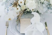 a chic wedding place setting with a clear charger, a white napkin and a fantastic wedding centerpiece of white blooms, gilded fronds and pale blue leaves