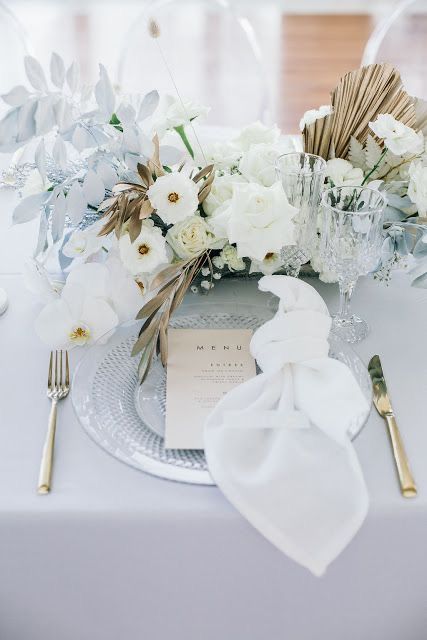 a chic wedding place setting with a clear charger, a white napkin and a fantastic wedding centerpiece of white blooms, gilded fronds and pale blue leaves