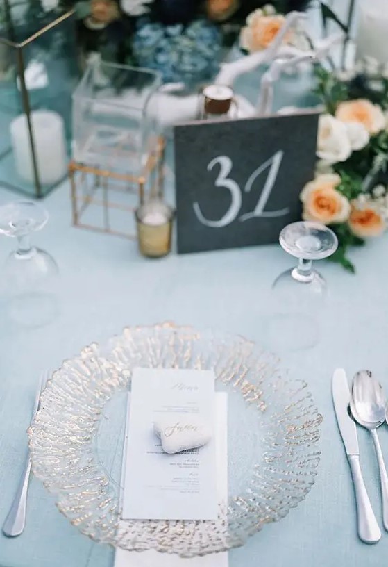 a chic wedding table setting with a pale blue tablecloth, a gilded plate, peachy blooms and neutral napkins