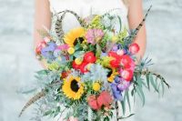 a colorful boho wedding bouquet with feathers, sunflowers, billy balls, greenery and red and purple blooms is cool for a boho bride