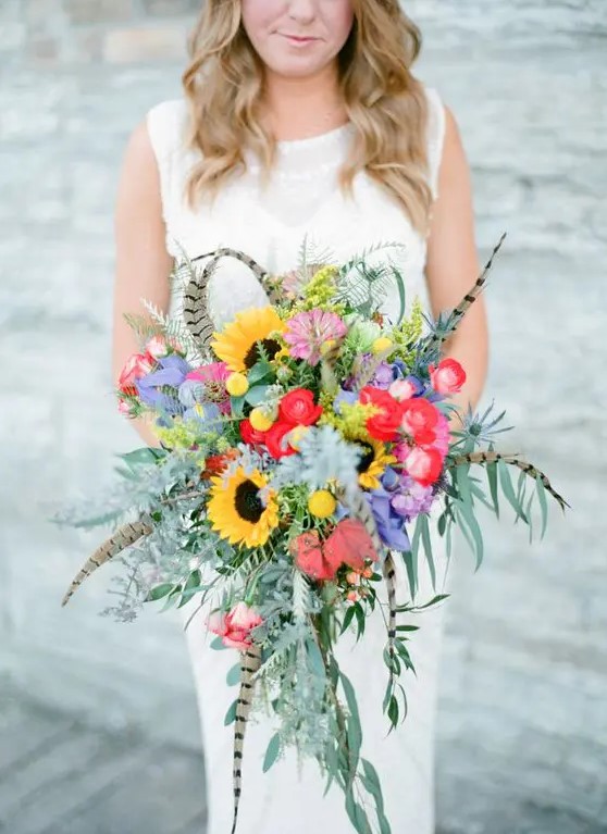 a colorful boho wedding bouquet with feathers, sunflowers, billy balls, greenery and red and purple blooms is cool for a boho bride