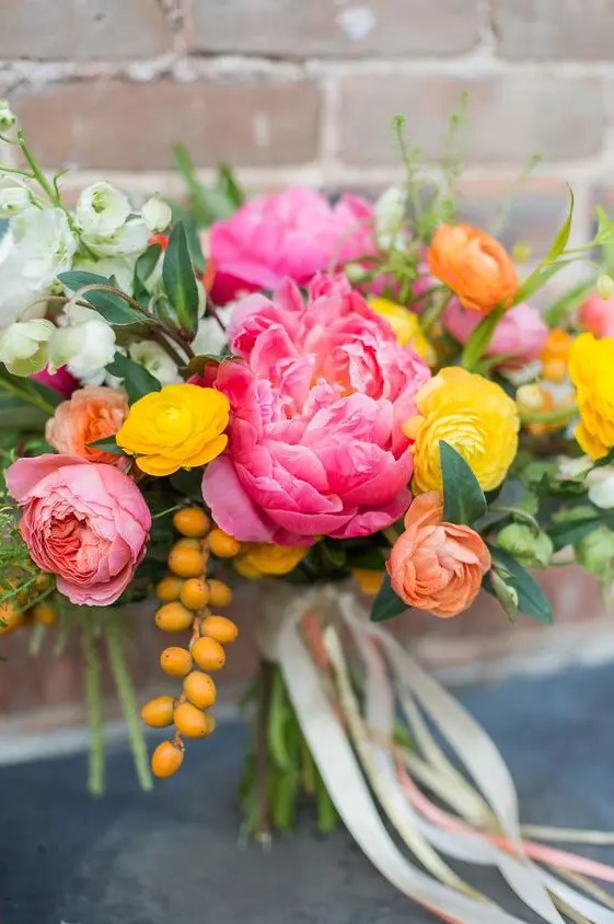 a colorful wedding bouquet with bright pink peonies, yellow and orange ranunculus, berries and white blooms and lots of greenery