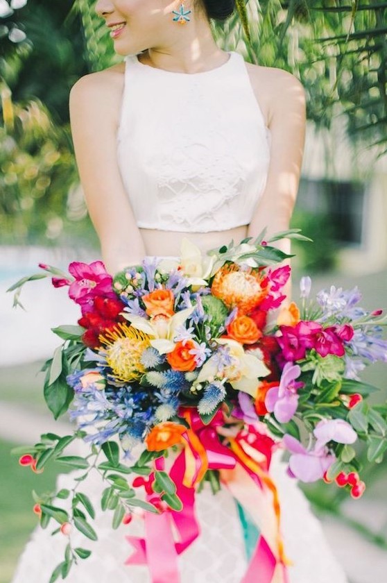 a colorful wedding bouquet with orange, fuchsia, pink, yellow and blue blooms, pincushion proteas and greenery plus colorful ribbons