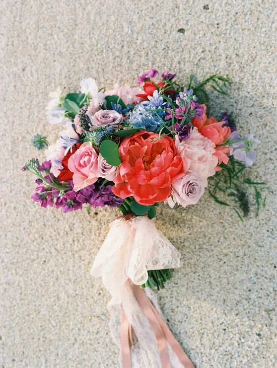a cool colorful wedding bouquet in red, mauve, pink, blue and purple plus lace and blush ribbons hanging down