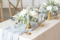 a delicate wedding tablescape with pale blue candles and a table runner, white blooms and greenery and gold vases and candleholders