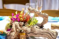 a driftwood wedding centerpiece with bright tropical blooms and candles in colorful glass candle holders