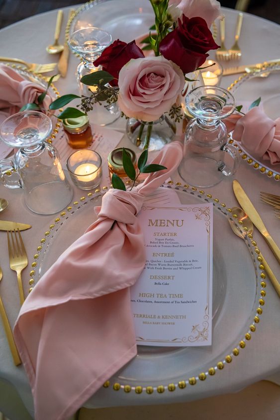 a glam and chic wedding tablescape with pink napkins and roses, gold cutlery and gold-rimmed chargers plus candles