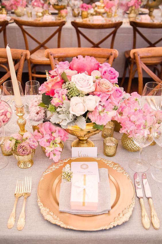 a glam and refined wedding tablescape with a bright pink floral centerpiece, a gold charger and cutlery, gold candleholders and vases