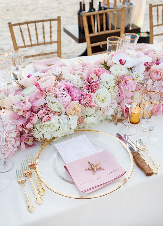 a glam beach wedding reception table with a lush light and bright pink centerpiece, a clear charger and a pink napkin, gold cutlery and gold-rimmed glasses