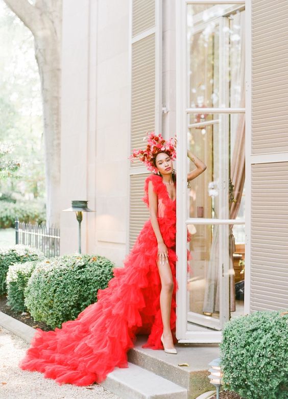 a hot red A-line wedding dress with a deep neckline, thick straps and a ruffle skirt with a train, a red floral crown and nude shoes for a bold and unique look
