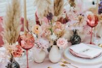 a modern wedding table setting with a blush tablecloth and napkins, gold cutlery, pink glasses and blooms and pampas grass