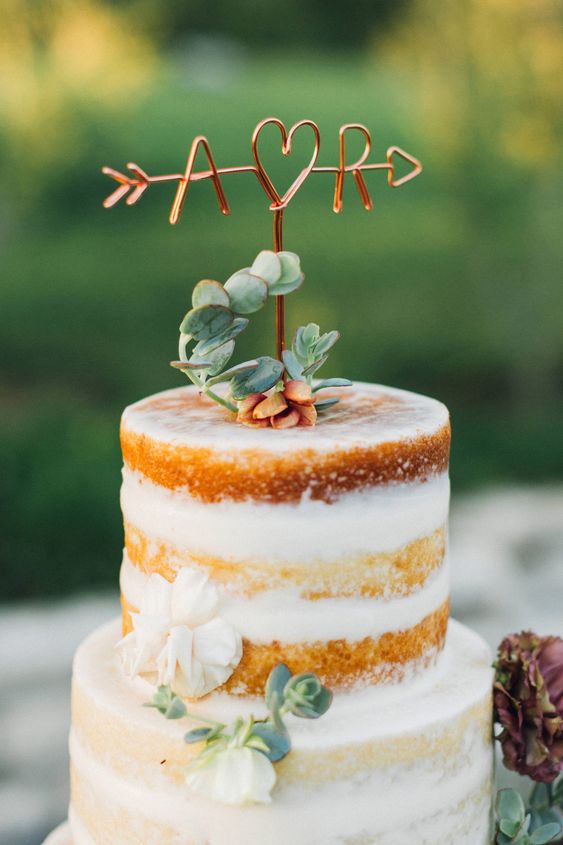 a naked wedding cake decorated with greenery and white blooms, with succulents and a copper wire monogram cake topper