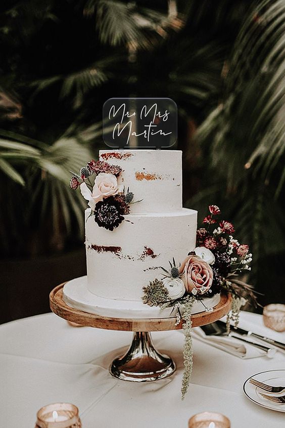 a naked wedding cake decorated with neutral and dark blooms, greenery and thistles, an acrylic and calligraphy cake topper for a modern wedding