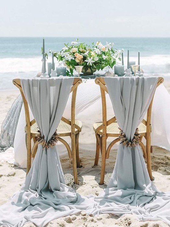 a pale blue beach wedding tablescape with light blue candles, glasses, plates and linens, neutral blooms and driftwood