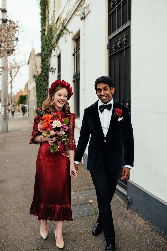 a refined and beautiful burgundy midi length wedding dress with a lace bodice, a shiny skirt and a ruffle trim, short sleeves and a red floral crown