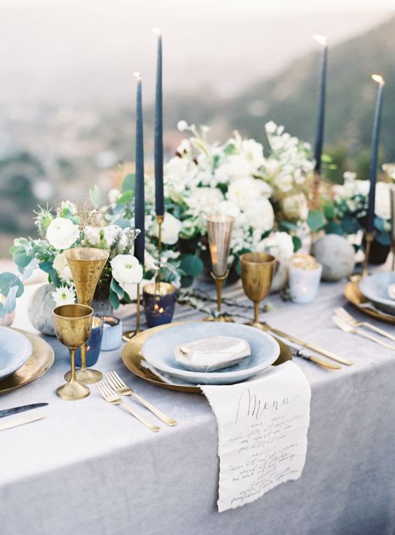 a refined pale blue wedding table setting with navy candles, pale blue plates and a tablecloth, gold goblets and white blooms