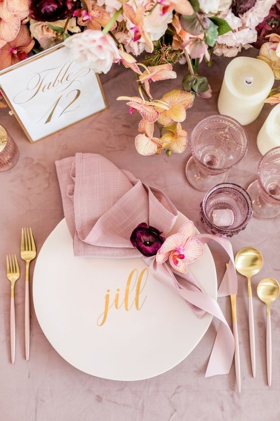 a sophisticated light pink and gold wedding table setting with light pink and mauve glasses, a light pink napkin and a tablecloth, gold cutlery