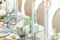 a stylish pale blue wedding tablescape with pale linens and glasses, white roses, greenery, neutral candles and gold cutlery