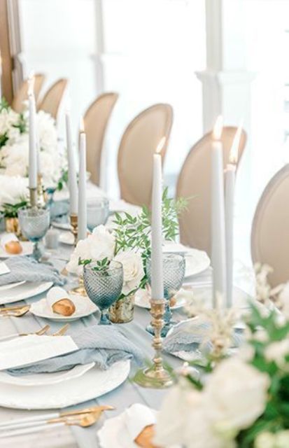a stylish pale blue wedding tablescape with pale linens and glasses, white roses, greenery, neutral candles and gold cutlery