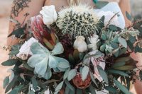 a stylish wedding bouquet of greenery, white and blush roses, succulents and a large cactus in the center