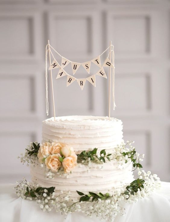 a white buttercream wedding cake decorated with greenery and blooms and with a banner cake topper is a lovely idea