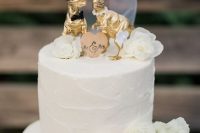 a white buttercream wedding cake decorated with white blooms, a wooden heart and fun gilded dino cake toppers dressed like a marrying couple