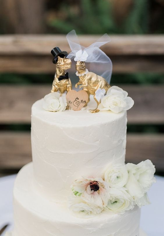 a white buttercream wedding cake decorated with white blooms, a wooden heart and fun gilded dino cake toppers dressed like a marrying couple